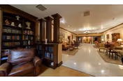 A spacious and warmly lit common area in a senior living facility featuring comfortable leather armchairs, wooden bookshelves filled with books and decorative items, and multiple seating arrangements with tables and chairs. The room has a polished floor, soft lighting from ceiling fixtures, and a cozy, inviting atmosphere.