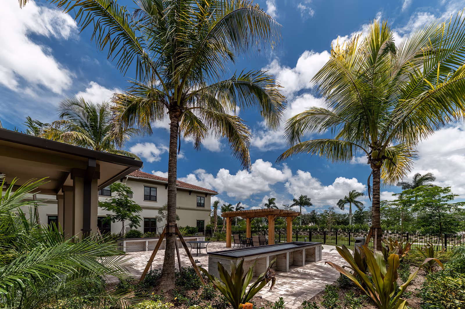 Outdoor patio area at The Capstone at Royal Palm Senior Living featuring palm trees, a wooden pergola with seating, landscaped garden beds, and a two-story building in the background under a partly cloudy blue sky.