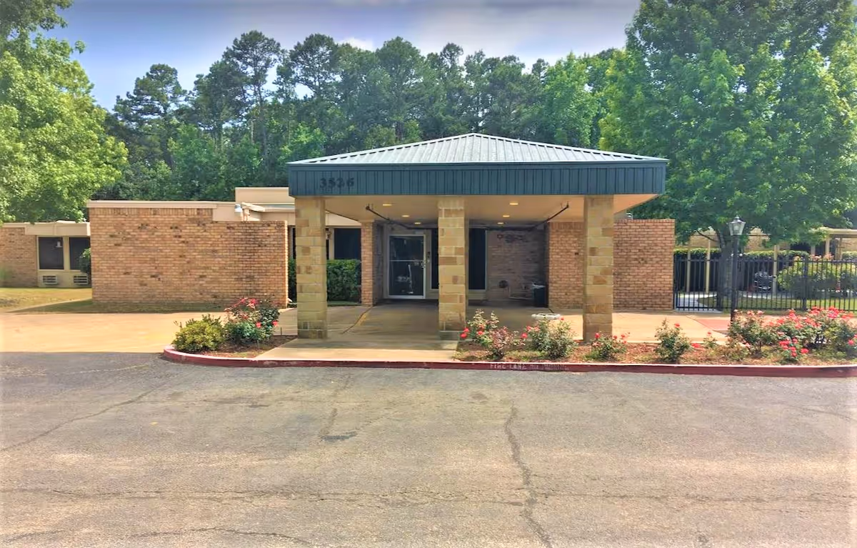 Front entrance of Greenbrier Nursing and Rehabilitation Center of Tyler showing a covered drop-off area with brick pillars, surrounded by landscaped bushes and flowers, with trees in the background.