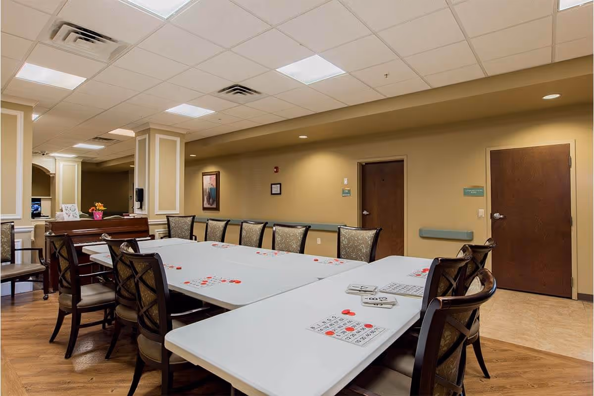 Interior view of a common area in a senior living facility with a long table set up for bingo, including bingo cards and red markers on the table. Several chairs are arranged around the table, and the room has beige walls, wooden doors, and a ceiling with recessed lighting.
