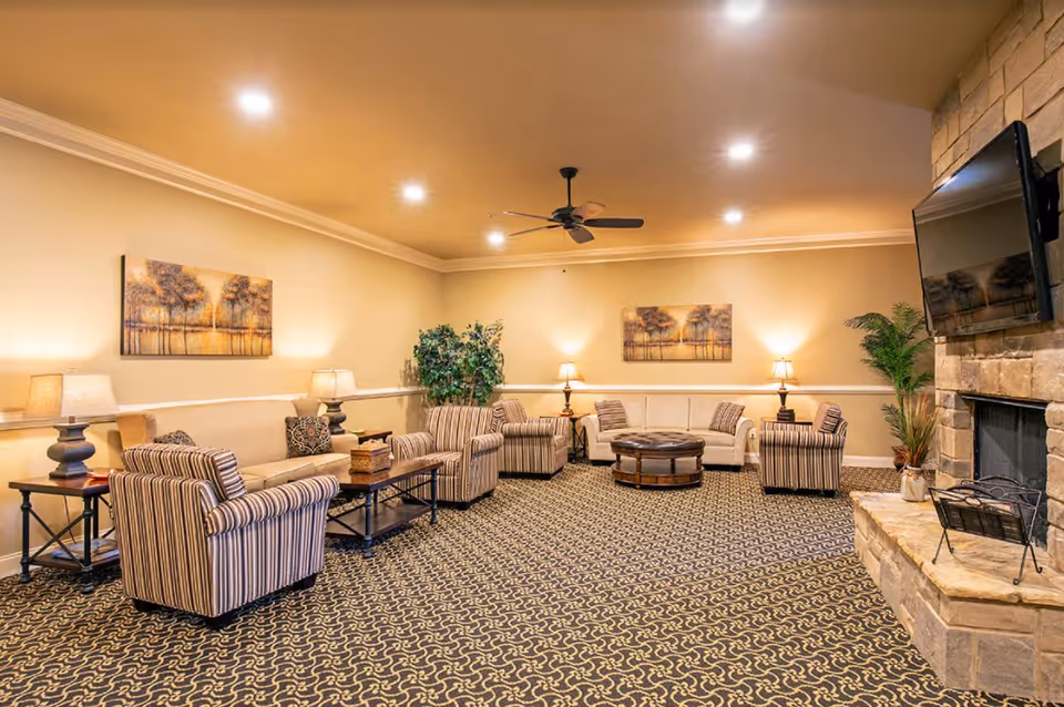 A spacious living room with patterned carpet, beige walls, and recessed ceiling lights. The room features multiple striped and solid beige armchairs and sofas arranged around a central round ottoman. There are two framed landscape paintings on the walls, several table lamps, a ceiling fan, a large stone fireplace with a mounted flat-screen TV, and some potted plants.