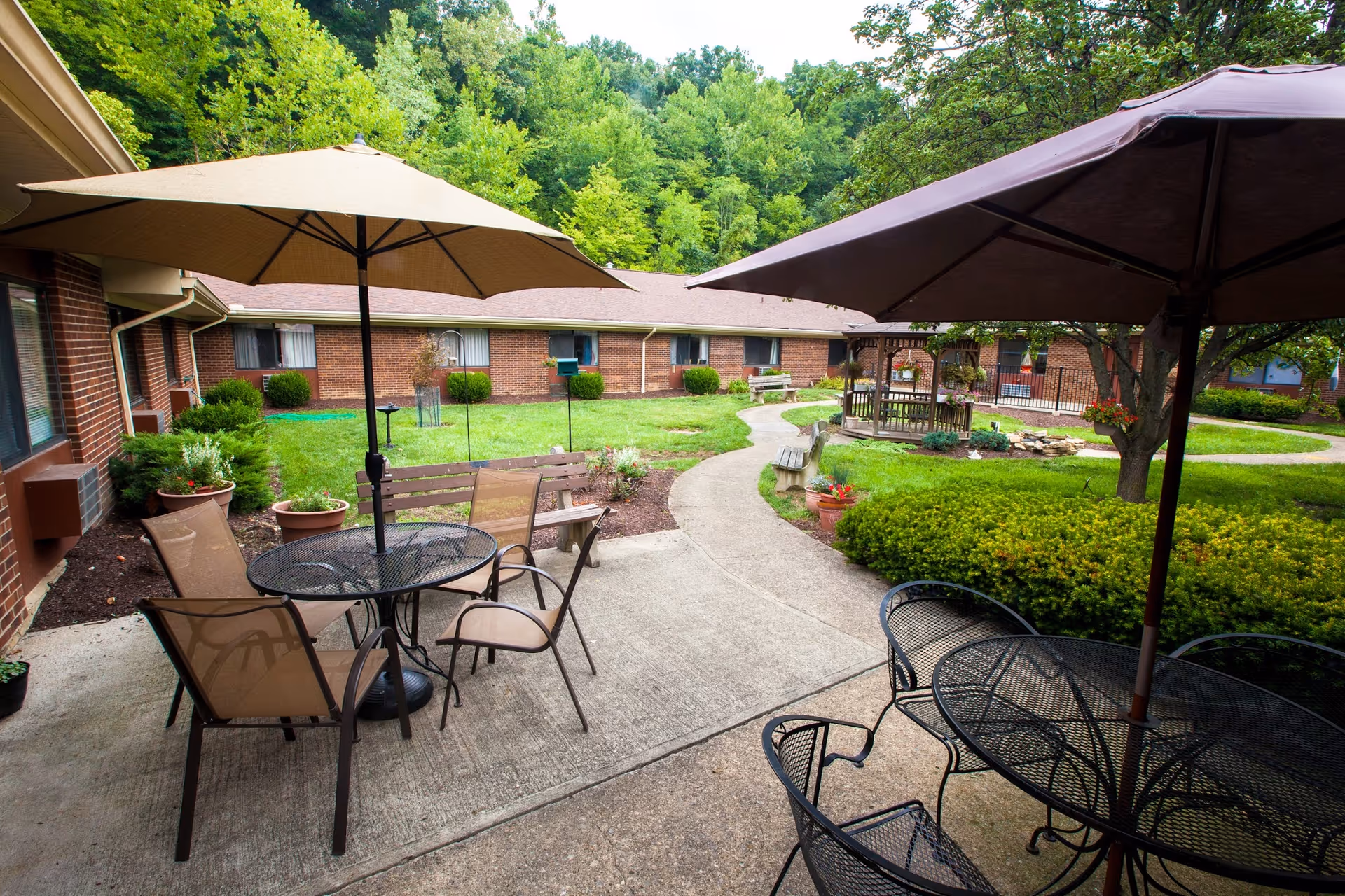 Outdoor courtyard area at Loveland Health Care featuring patio tables with umbrellas, chairs, a winding concrete pathway, green bushes, a small gazebo, and a backdrop of trees and a brick building.