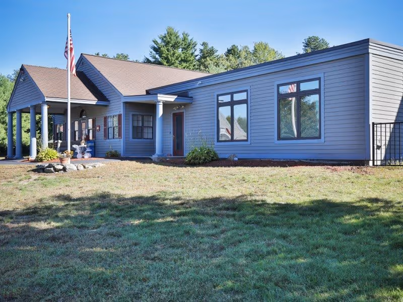 Exterior view of a single-story building with gray siding and a brown roof, featuring several windows and a front entrance with columns. An American flag is displayed on a flagpole in front of the building, surrounded by a small landscaped area with plants and rocks. The building is set against a backdrop of trees and a clear blue sky.