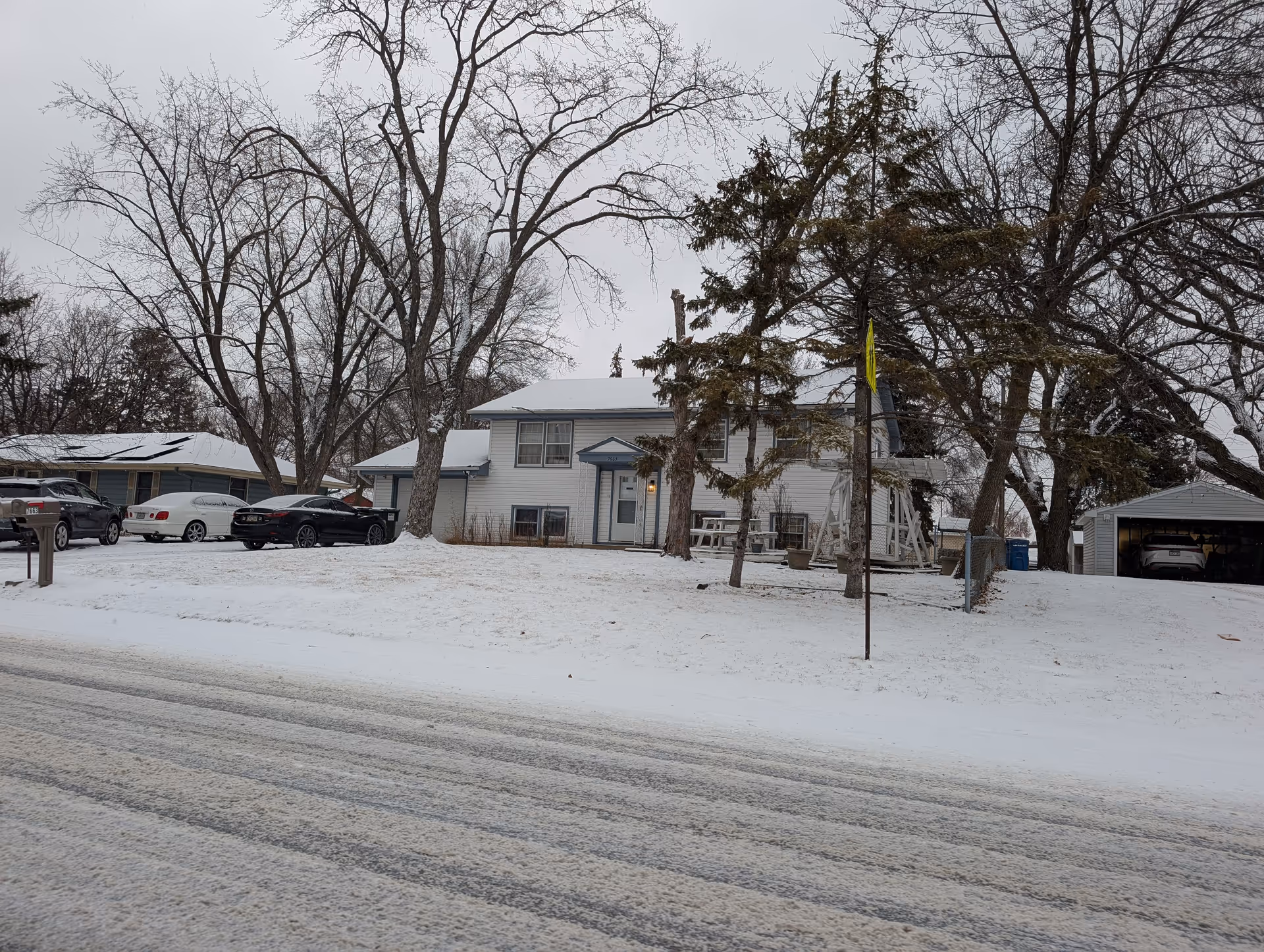 A snowy residential street with a white two-story house in the center. The house has a small porch with a light on, several leafless trees around it, and a detached garage to the right with a car inside. There are multiple cars parked along the street and driveway. The sky is overcast.