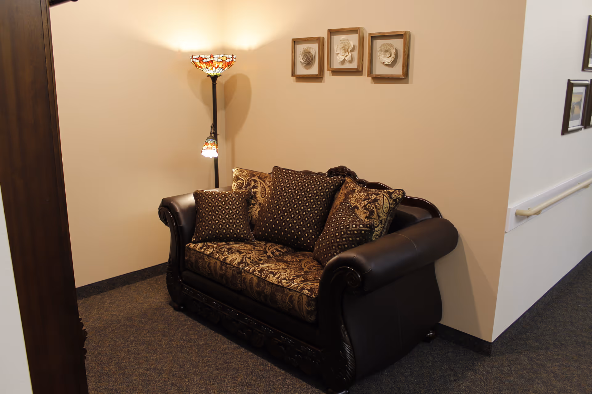 A cozy corner with a dark ornate loveseat featuring patterned cushions, a Tiffany-style floor lamp with stained glass shades, and three framed floral artworks on a beige wall in a carpeted hallway.