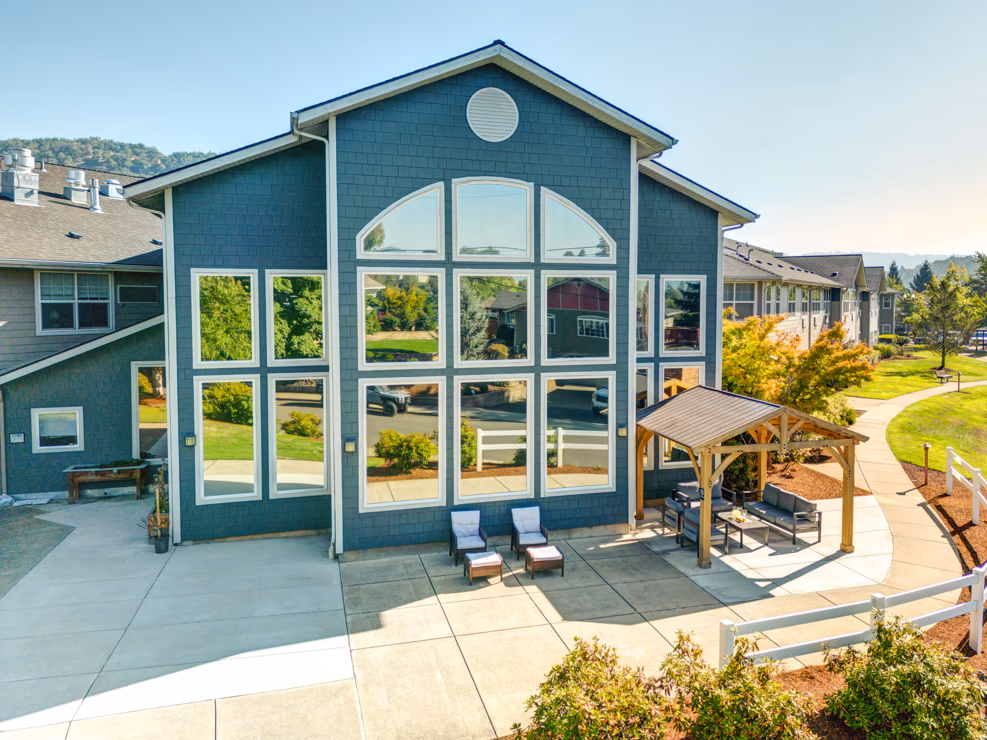 Exterior view of a modern retirement and assisted living facility building with large windows reflecting the surrounding greenery. There is a small outdoor seating area with two lounge chairs and a covered patio with additional seating. The building is surrounded by landscaped pathways and trees under a clear sky.