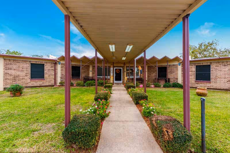 Covered walkway leading to the entrance of a single-story brick building with a well-maintained lawn and flower beds on either side under a blue sky.