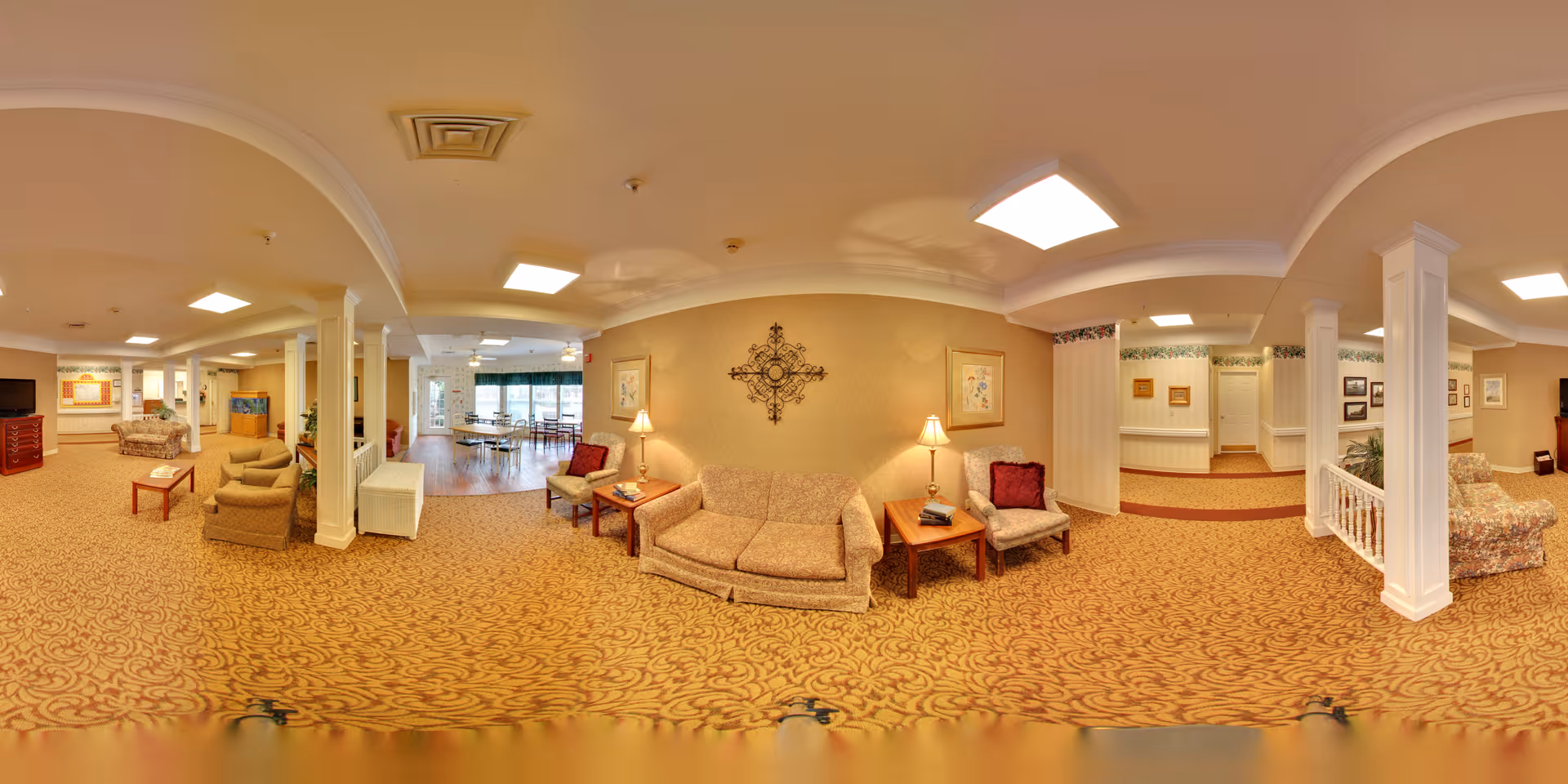 A spacious and warmly decorated senior living facility common area with patterned carpet, multiple seating arrangements including sofas and armchairs, side tables with lamps, framed artwork on the walls, and a dining area with tables and chairs visible in the background near large windows.