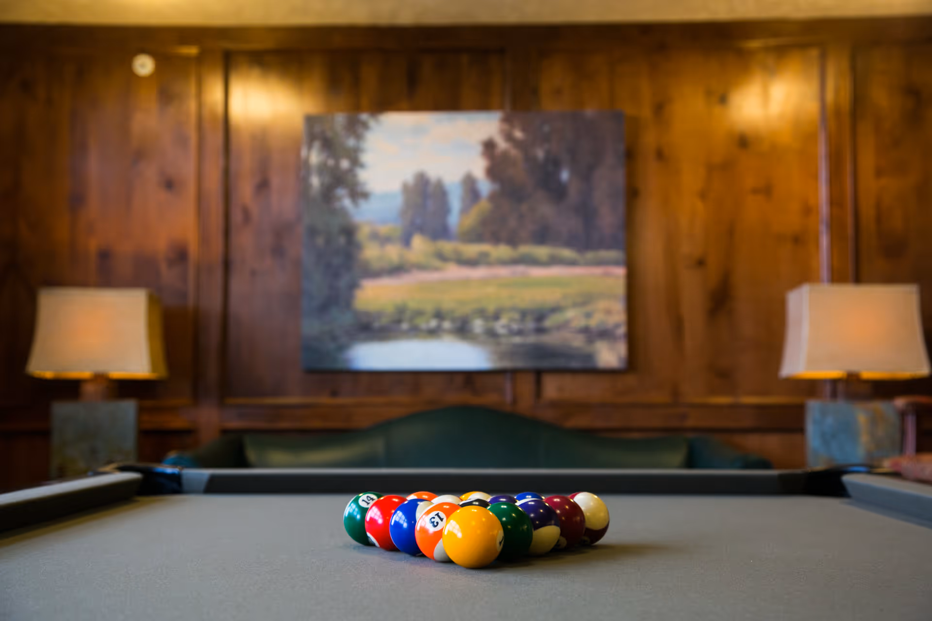A pool table with a rack of colorful billiard balls arranged in the center. The background features a wooden paneled wall with a landscape painting and two table lamps on either side of a green couch.