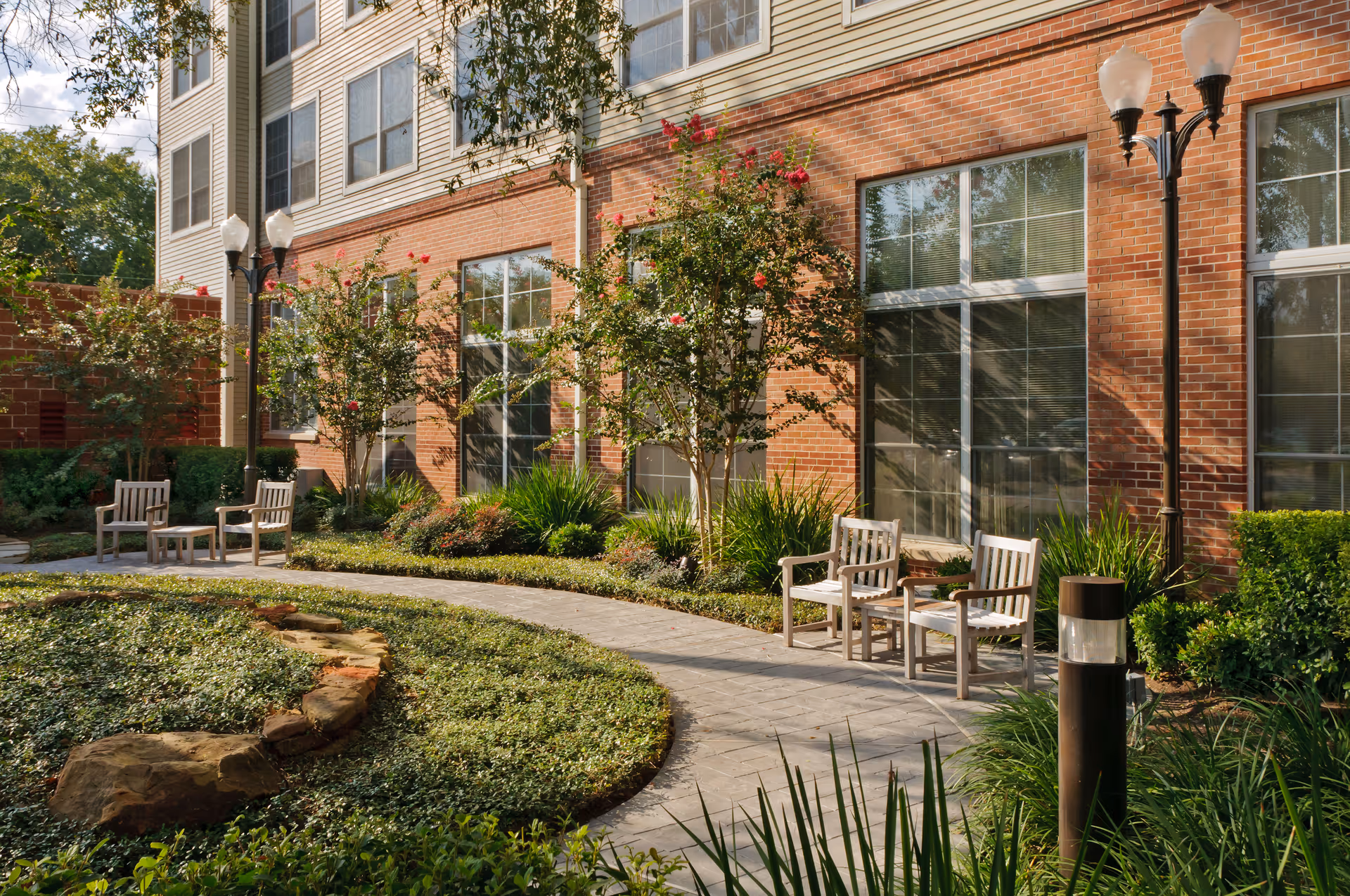 Outdoor garden area at The Vosswood Nursing Center featuring a curved paved walkway, green shrubs, flowering trees, and wooden chairs arranged in pairs along the path next to a brick building with large windows and lamp posts.