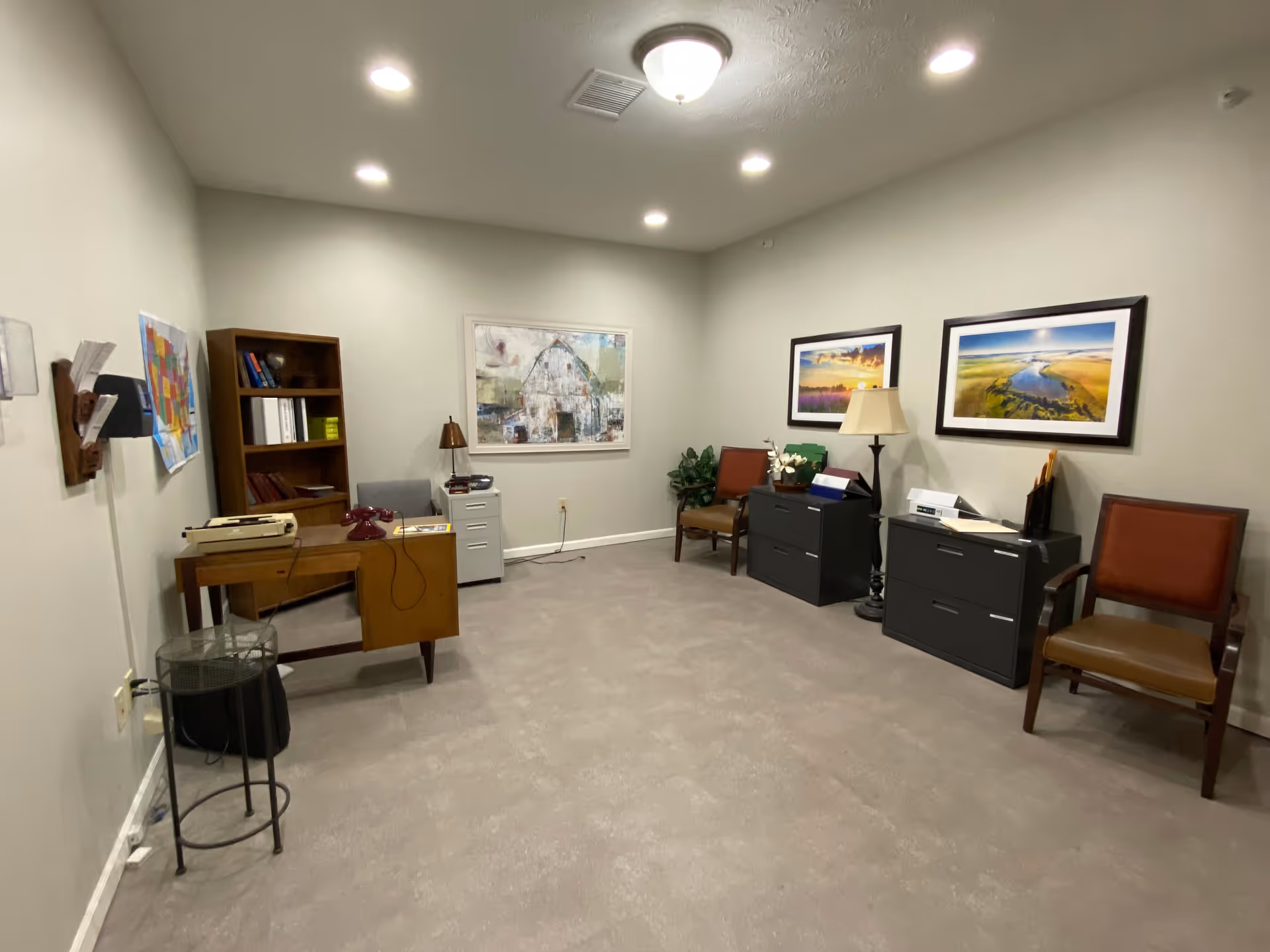 Small office-style interior with desks, filing cabinets, chairs, a bookshelf, and framed artwork on the walls.