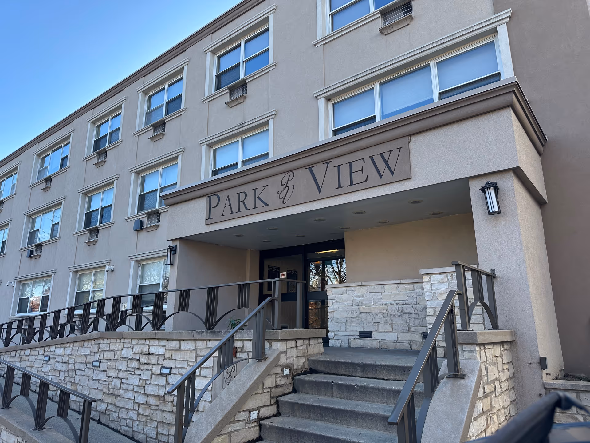 Exterior front entrance of a multi-story building with a sign above the entrance reading 'PARK VIEW'. The building has multiple windows with air conditioning units and a stone and metal railing leading up to the entrance steps.