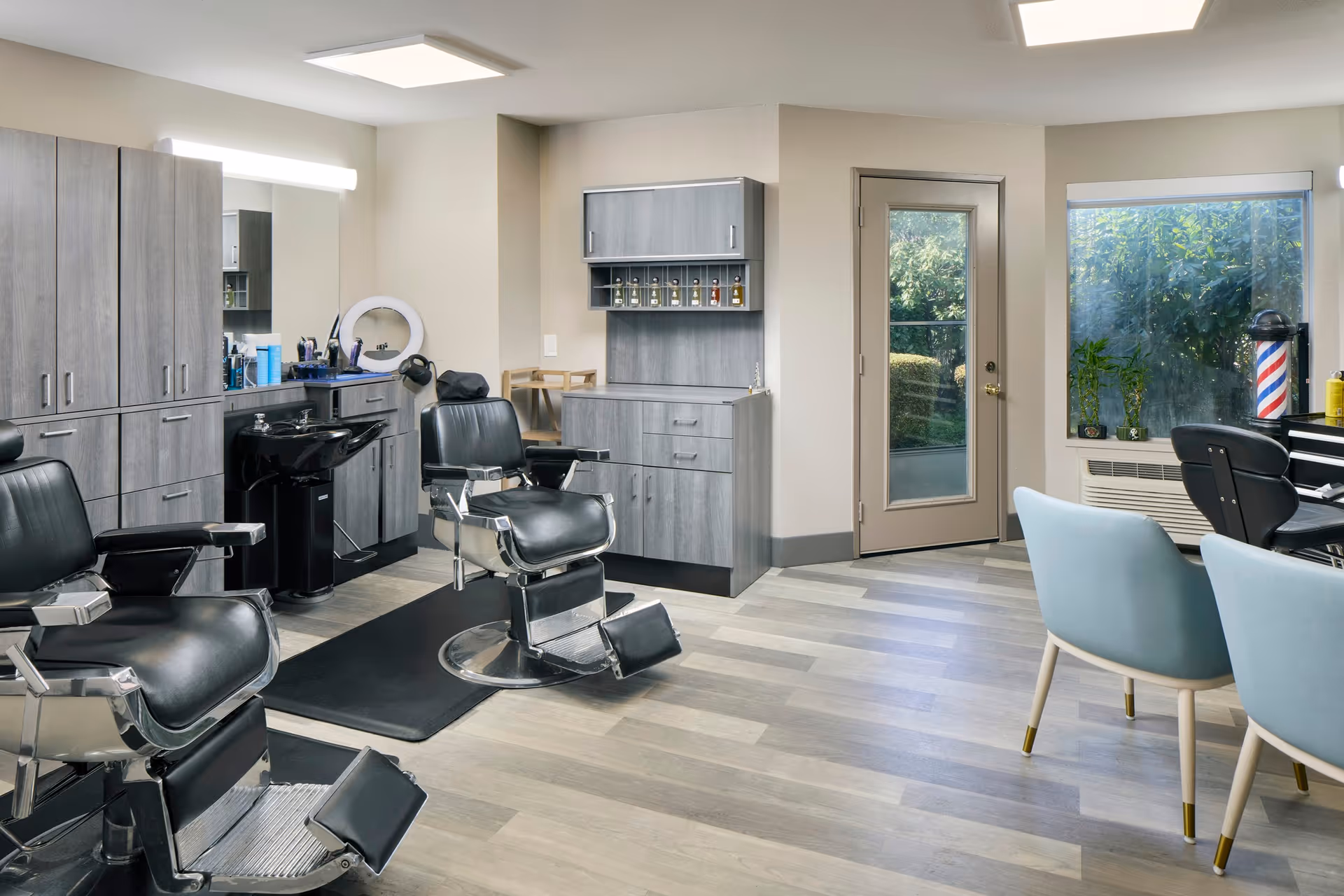 Interior salon/barber room with black barber chairs, shampoo sink, gray cabinetry, and seating by a window and door.