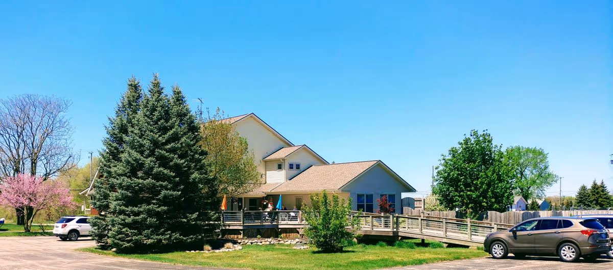 Front exterior of a two-story cottage-style senior living building with a wooden ramp, lawn, trees, and parked cars under a clear blue sky.