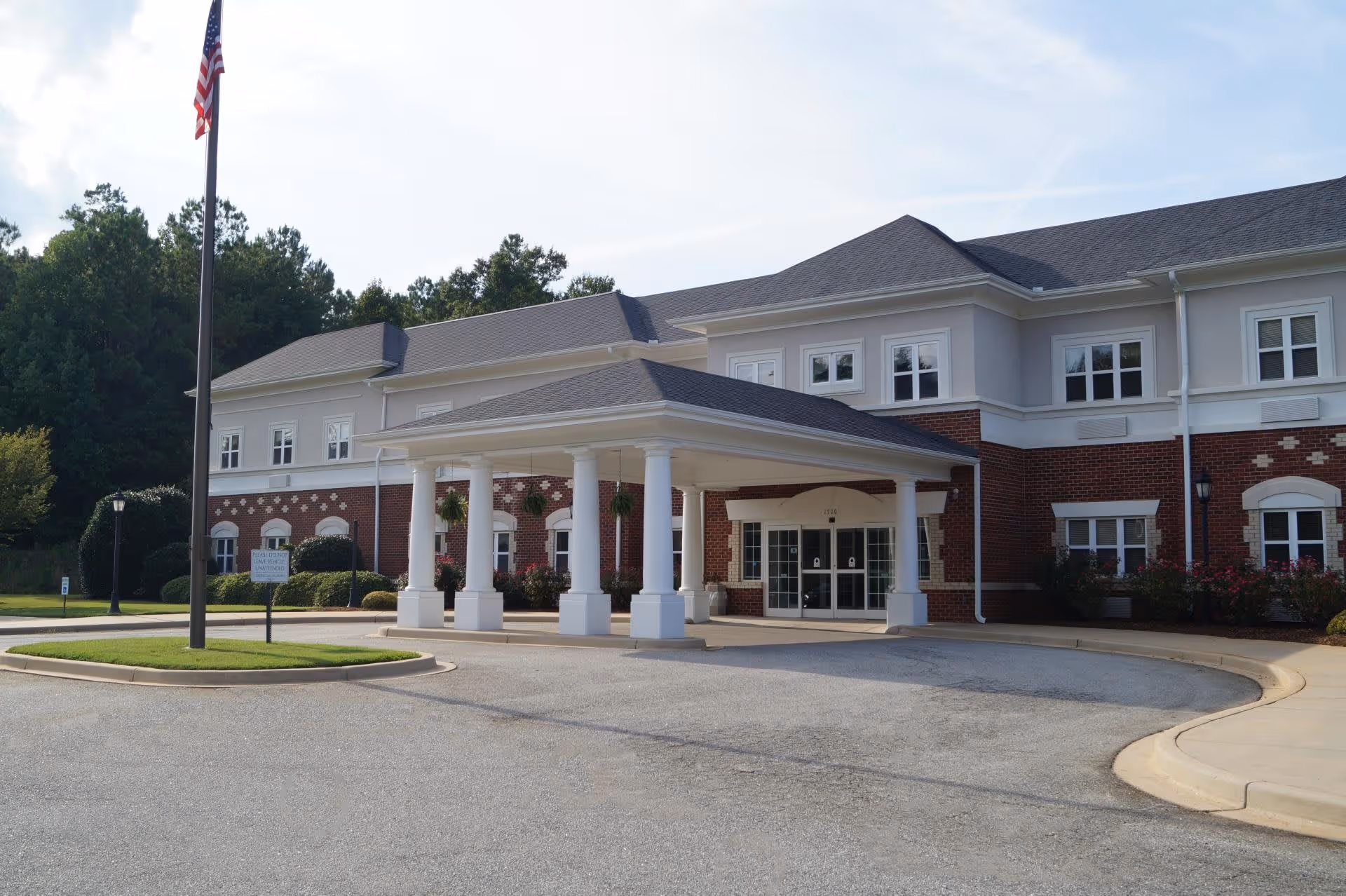 Exterior view of a two-story senior living facility building with a covered entrance supported by white columns. The building has a combination of red brick and light-colored siding, multiple windows, and a flagpole with an American flag in front. There are landscaped bushes and a paved driveway leading to the entrance.