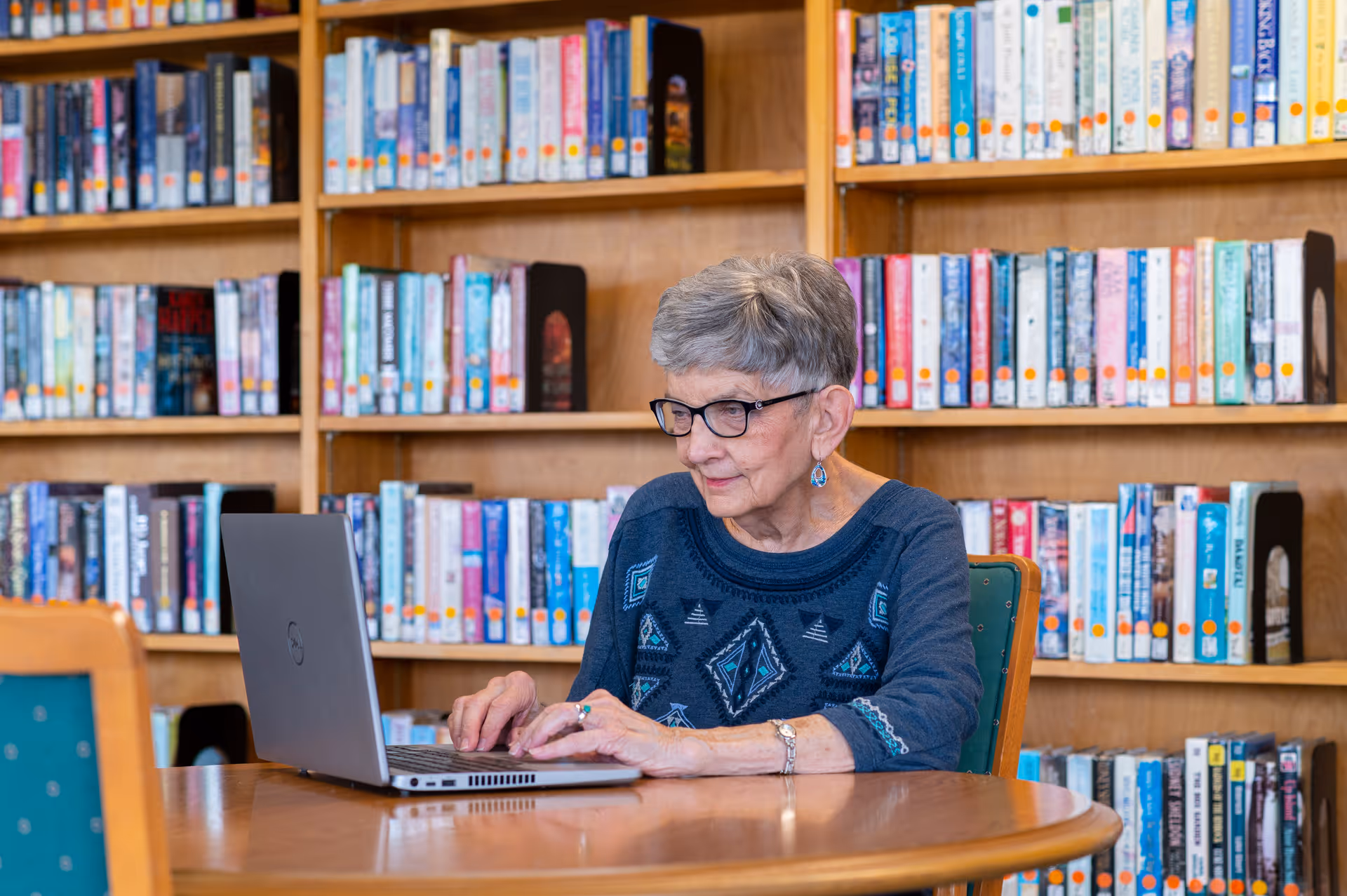 An elderly woman with short gray hair and glasses is sitting at a round wooden table using a laptop. Behind her are wooden bookshelves filled with books, indicating a library or reading area.