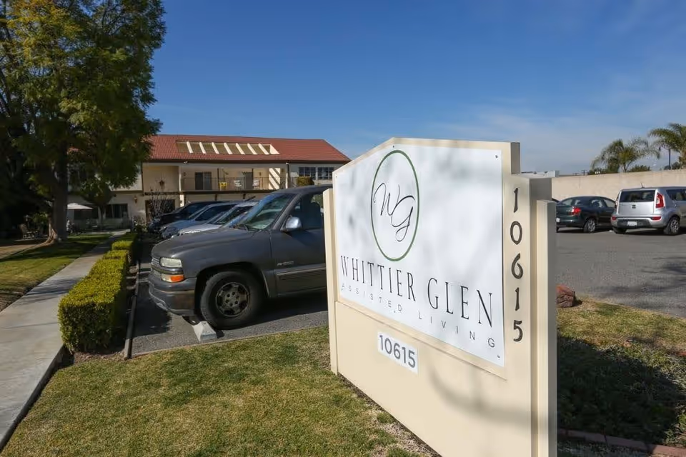 Outdoor view of Whittier Glen Assisted Living facility sign with the address 10615, parked cars, a sidewalk, and a building with a red roof in the background under a clear blue sky.