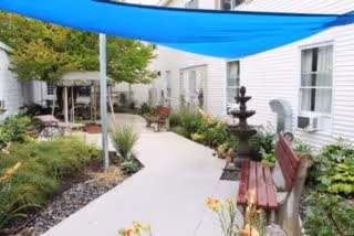 Outdoor courtyard area at Memory Care at Market Square featuring a concrete walkway, benches, a multi-tiered water fountain, plants, and a blue shade canopy overhead. The courtyard is surrounded by white building walls with windows and air conditioning units.