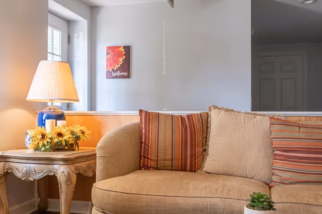 Cozy living room area with a beige couch adorned with striped and plain cushions. Next to the couch is an ornate wooden side table holding a blue lamp with a beige lampshade and a small bouquet of yellow sunflowers. A window lets in natural light, and a small wall decoration with a sunflower and the word 'Welcome' hangs on the wall.