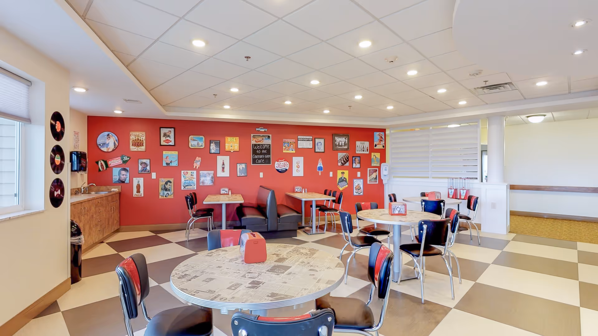 A bright and colorful dining area with retro-style round tables and chairs. The floor has a checkered pattern in beige and brown. One wall is painted red and decorated with vintage posters and signs, including a chalkboard sign that says 'Welcome to the Covenant Glen Cafe.' There is a window with blinds on the left and a counter with a sink beneath it. The ceiling has recessed lighting.