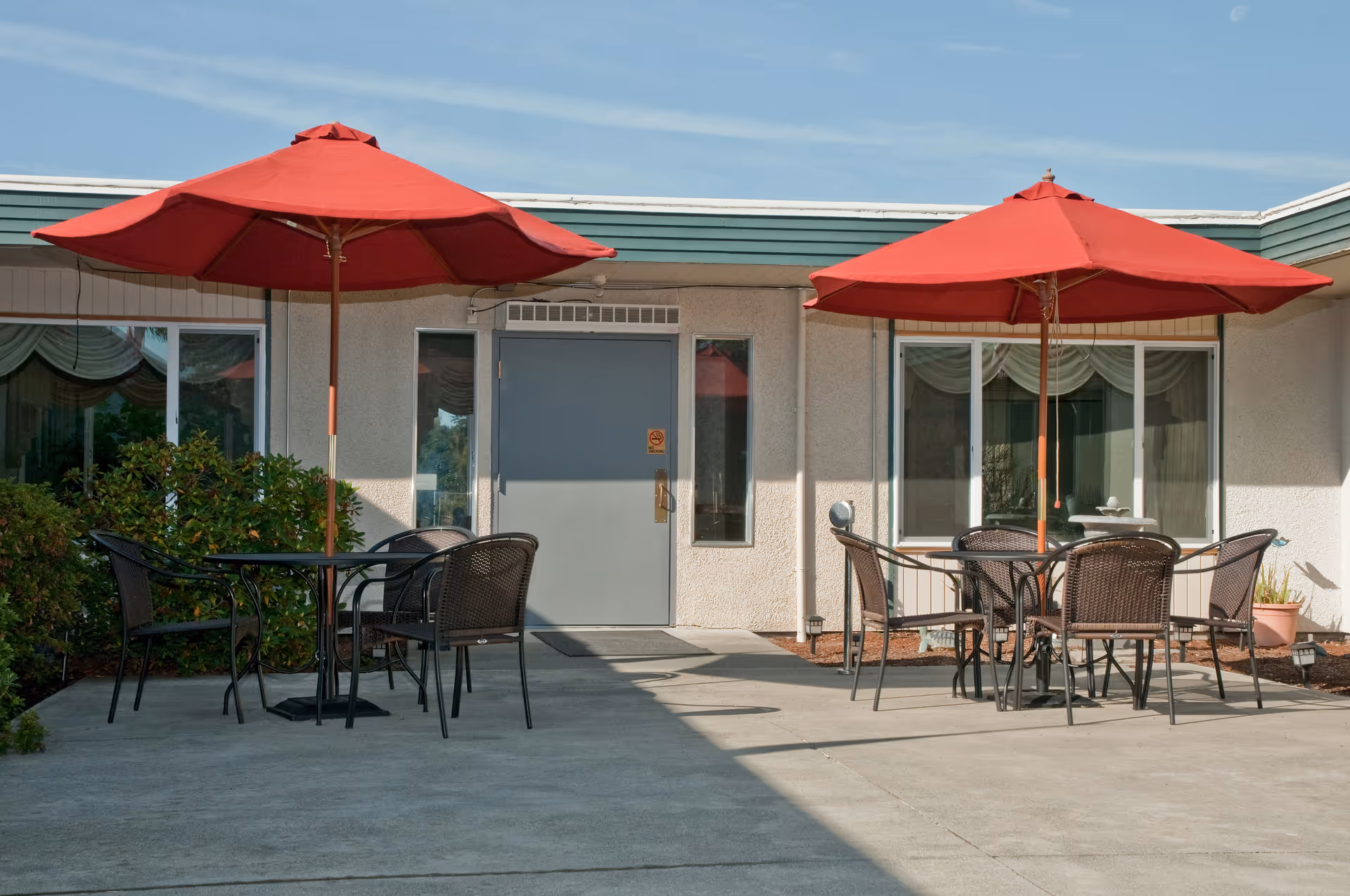 Outdoor patio area with two black metal tables, each shaded by a large red umbrella. Each table is surrounded by four black metal chairs. The patio is adjacent to a building with white walls, green trim, and windows with curtains. There is a gray door in the center of the building wall behind the tables.