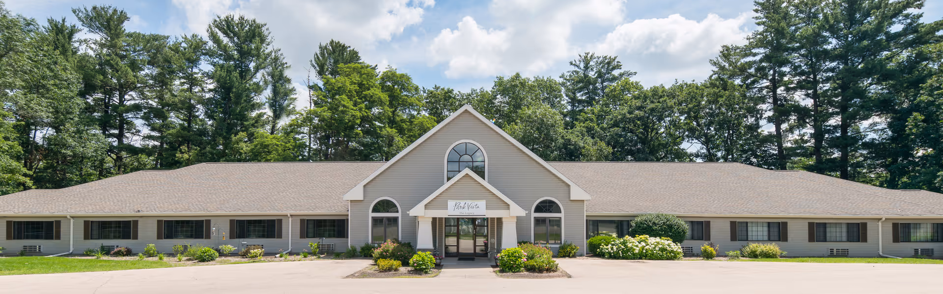 Front exterior view of a single-story senior living facility building named Park Vista The Legacy, surrounded by greenery and trees under a partly cloudy sky.