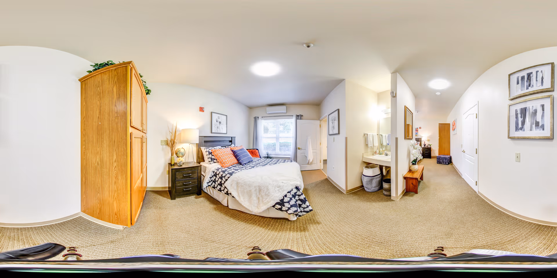 A spacious, well-lit bedroom in a senior living facility with a bed featuring patterned bedding and multiple pillows. There is a wooden wardrobe on the left, a nightstand with a lamp and globe beside the bed, and a window with curtains behind the bed. To the right, there is an open vanity area with a sink and mirror, and a hallway leading to other rooms. The walls are decorated with framed artwork, and the carpet is beige.