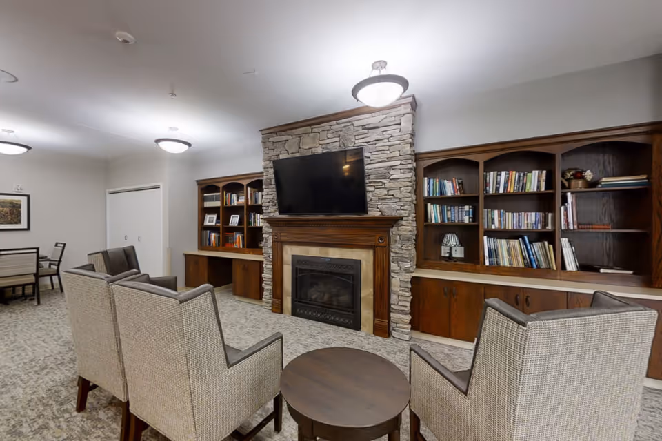 Cozy common living room with a stone fireplace, wall-mounted TV, built-in bookshelves and upholstered chairs around a small round table.