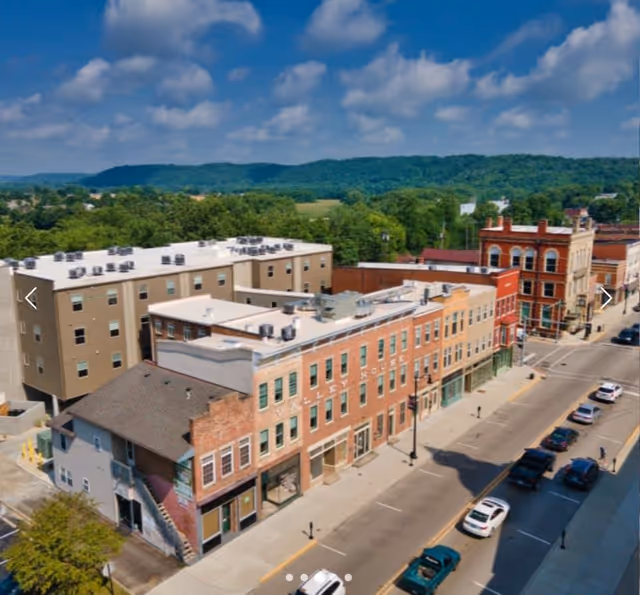 Aerial view of a small town street with multiple brick buildings and parked cars along the road. The background features green hills and a partly cloudy blue sky.
