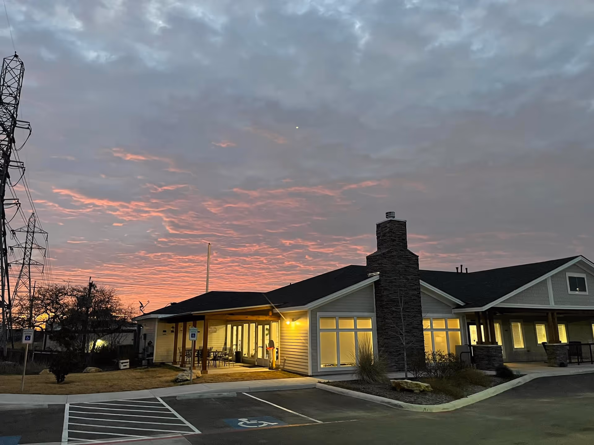 Exterior view of a single-story assisted living facility building at dusk with a dramatic cloudy sky showing pink and orange hues. The building has large windows with warm lights inside, a stone chimney, and a covered entrance with outdoor seating. There are parking spaces including handicapped spots in front of the building.