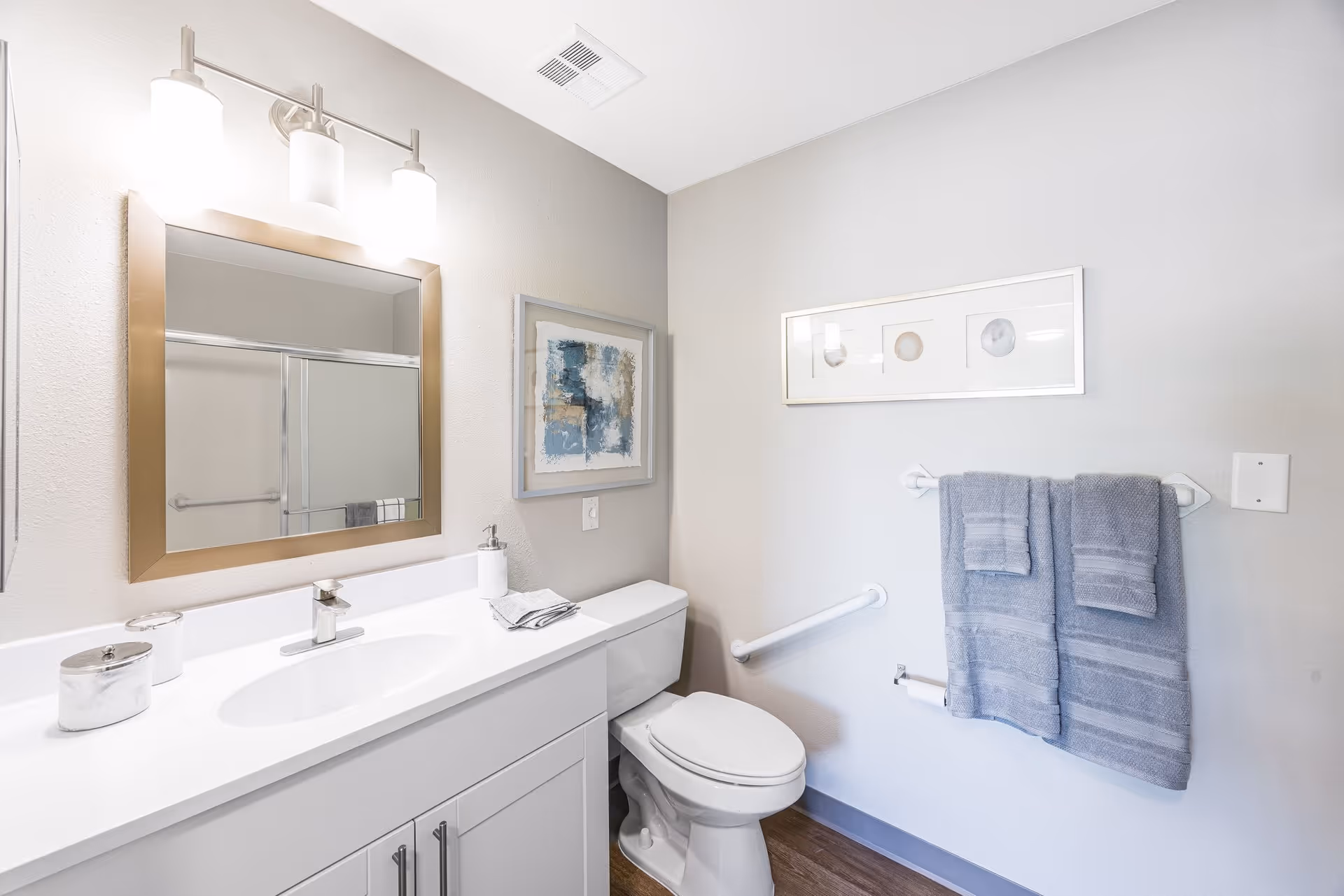 A clean and modern bathroom featuring a white vanity with a sink, a large mirror with a gold frame, and three light fixtures above it. There is a toilet next to the vanity, a towel rack with two gray towels, a grab bar on the wall, and framed artwork hanging above the toilet. The walls are painted light gray and the floor has wood-like flooring.