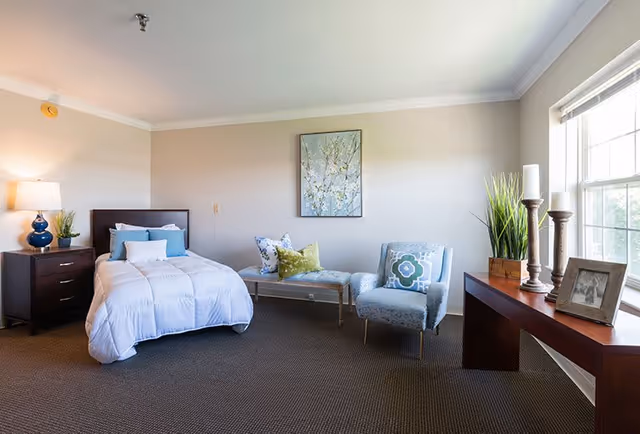 A bright and cozy bedroom in a senior living facility featuring a single bed with white bedding and blue pillows, a dark wooden nightstand with a blue lamp and a small plant, a cushioned bench with decorative pillows, a patterned armchair, a wooden console table with candles, a framed photo, and a potted plant, all set against beige walls and a large window letting in natural light.