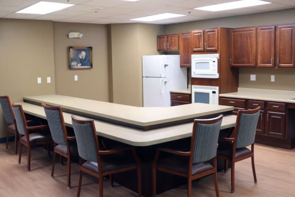A kitchen area with a U-shaped counter surrounded by several wooden chairs with cushioned seats and backs. The kitchen has dark wood cabinets, a white refrigerator, a microwave, and a built-in oven. The walls are beige, and there is a framed picture hanging on one wall.