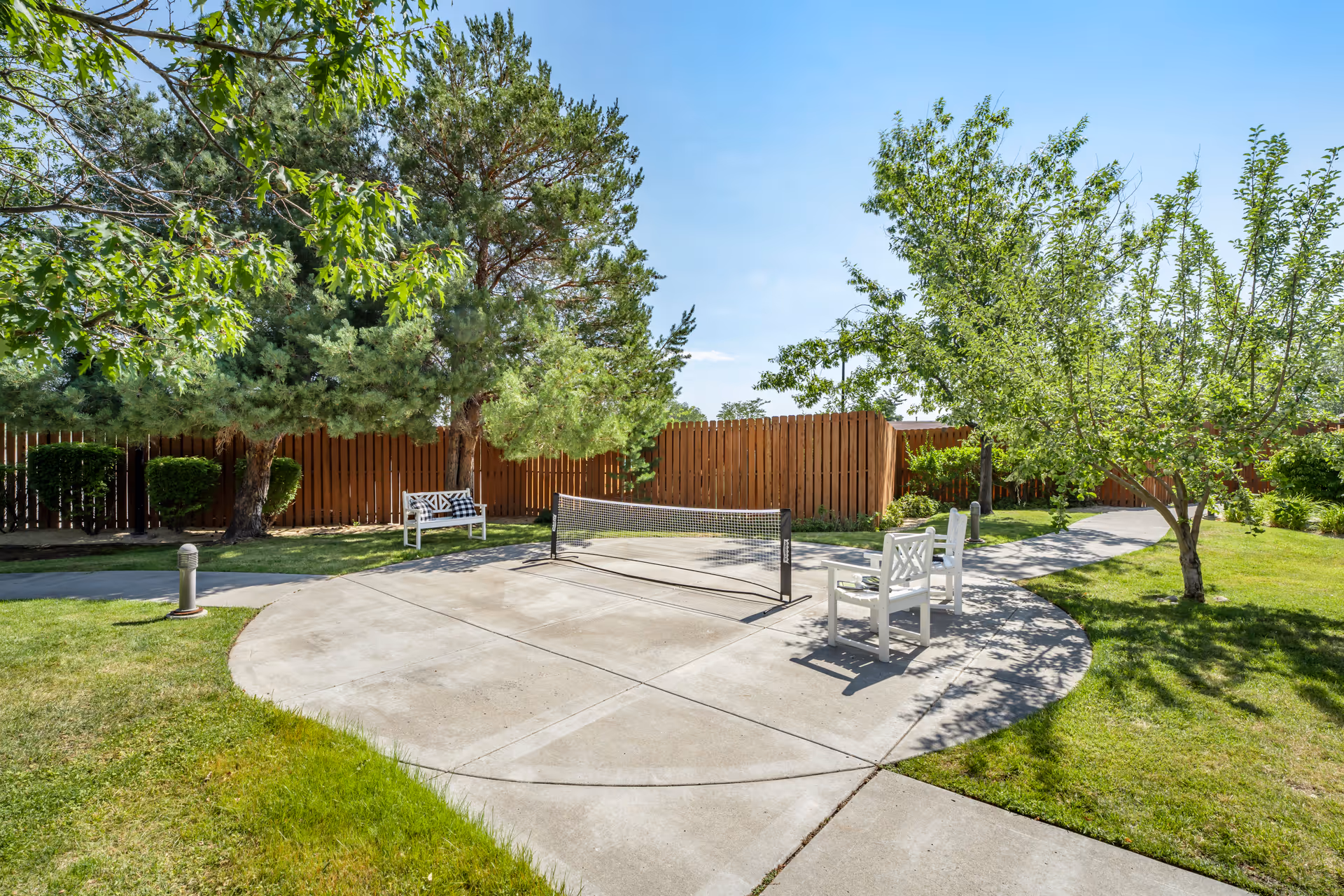 Outdoor recreational area with a small portable tennis or pickleball net set up on a circular concrete court surrounded by green grass and trees. There are white benches with cushions placed around the court, and a wooden fence encloses the area under a clear blue sky.