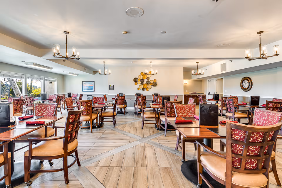 A spacious dining room with multiple wooden tables and chairs featuring red patterned cushions. The room is well-lit with natural light coming through large windows on the left side and several modern chandeliers hanging from the ceiling. The floor has a light wood tile design, and the walls are decorated with a circular metal wall art and a clock. Menus and red napkins are placed on the tables.