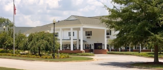 Front entrance of a two-story senior living facility with white columns, a covered portico, landscaped driveway, and an American flag.