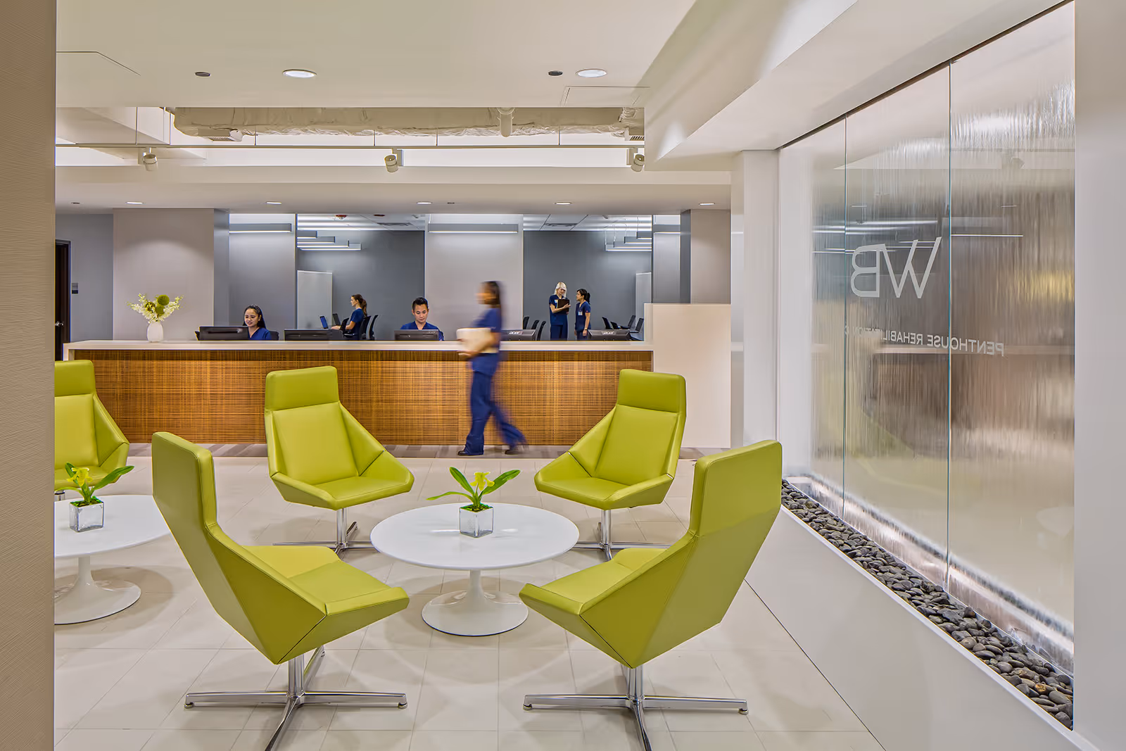 Modern reception area with a wooden front desk where three staff members are seated and one is walking. In the foreground, there are four bright green chairs arranged around a white circular table with a small plant. On the right side, there is a large glass wall with a water feature and the letters 'WB' along with the words 'PENTHOUSE REHABILITATION' visible through the glass.