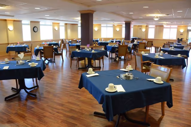 A spacious dining room with multiple tables covered in dark blue tablecloths, each set with white cups, saucers, napkins, and small centerpieces. The room has wooden flooring, several windows letting in natural light, and beige walls with wall-mounted lights and a clock.