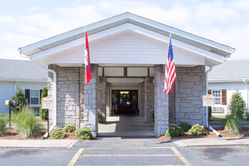 Covered stone entrance of a nursing facility with an American flag, another flag, and handicapped parking signs.