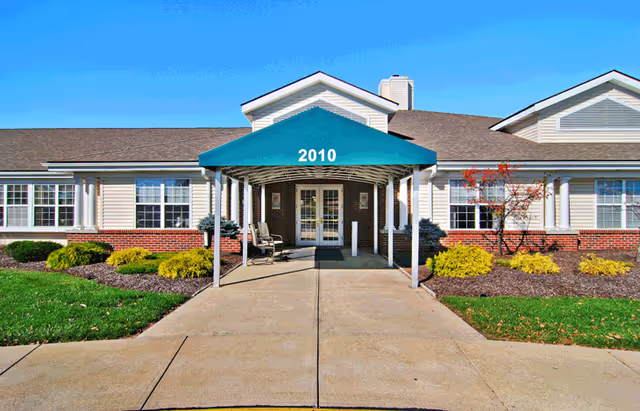 Front exterior view of a single-story senior living facility building with a green awning displaying the number 2010 above the entrance. The building has white siding, brick accents, multiple windows, and landscaped bushes and grass on either side of the concrete walkway leading to the entrance.