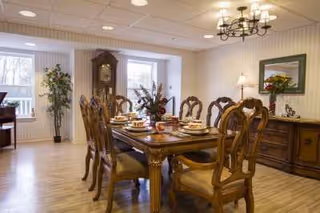 A well-lit dining room with a long wooden dining table set for six people with plates, cups, and utensils. The room features wooden chairs with cushioned seats, a wooden sideboard with a lamp and a framed picture above it, a chandelier hanging from the ceiling, and two windows letting in natural light. There is also a tall grandfather clock and a potted plant near the windows.