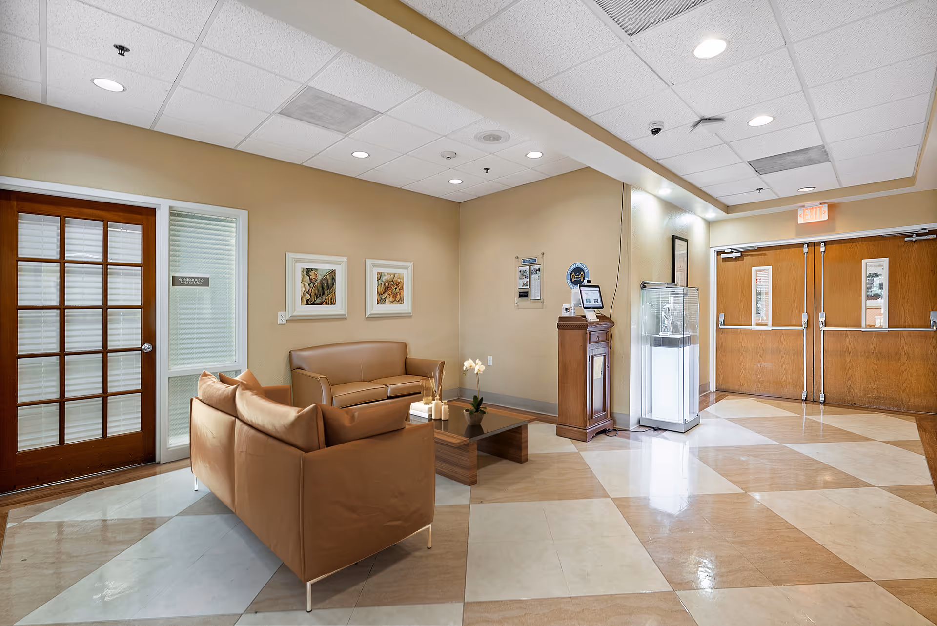 A well-lit interior common area with beige walls and a tiled floor featuring a diamond pattern. The space includes two tan leather sofas arranged around a wooden coffee table with decorative items. On the left, there is a wooden door with glass panels and a window with blinds. The back wall has two framed pictures. To the right, there is a wooden podium with a tablet on top and a glass display case next to it. Double wooden doors with small windows are visible in the background.