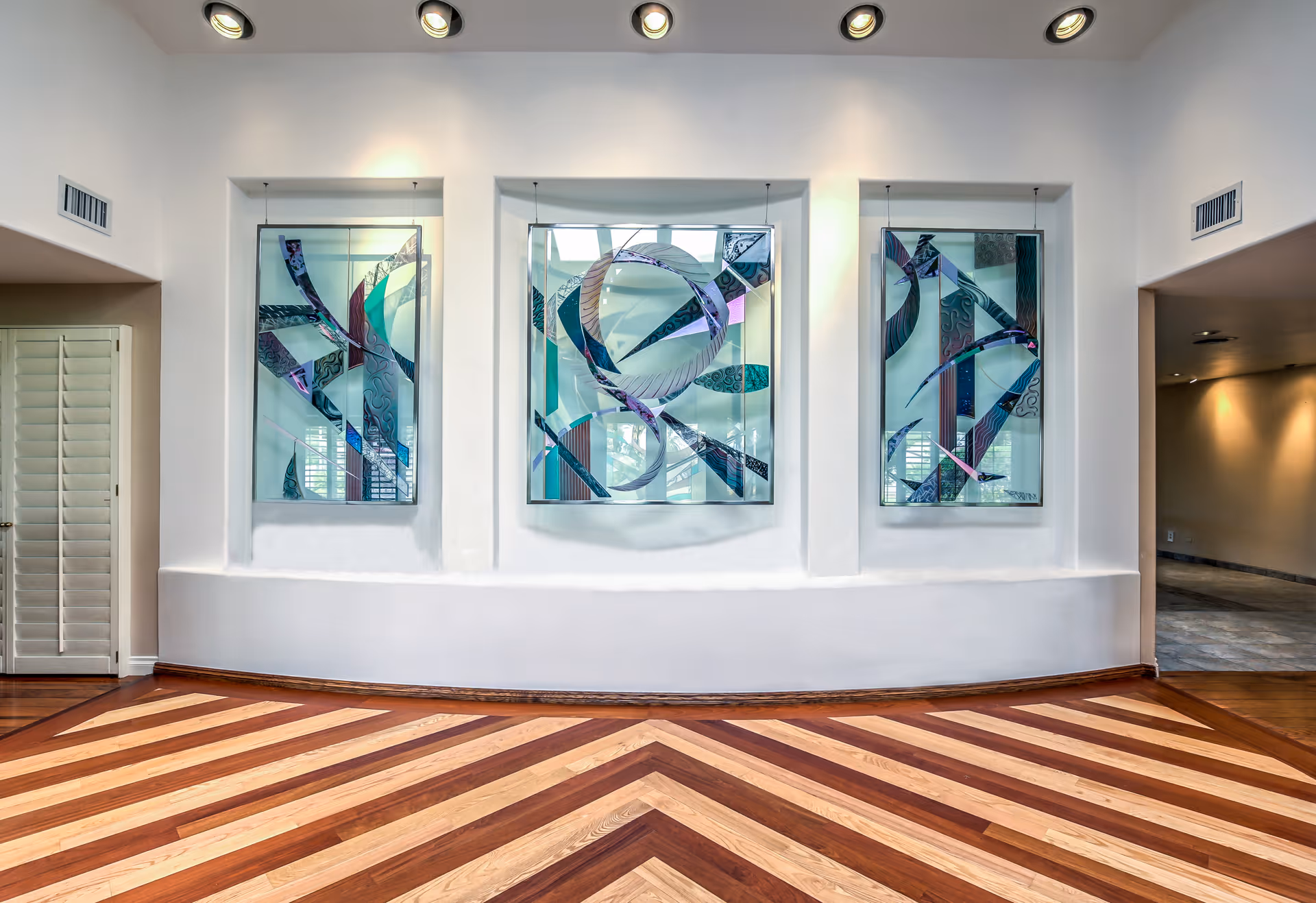 Interior common area with three decorative stained-glass panels set into a white wall above chevron-pattern hardwood flooring.