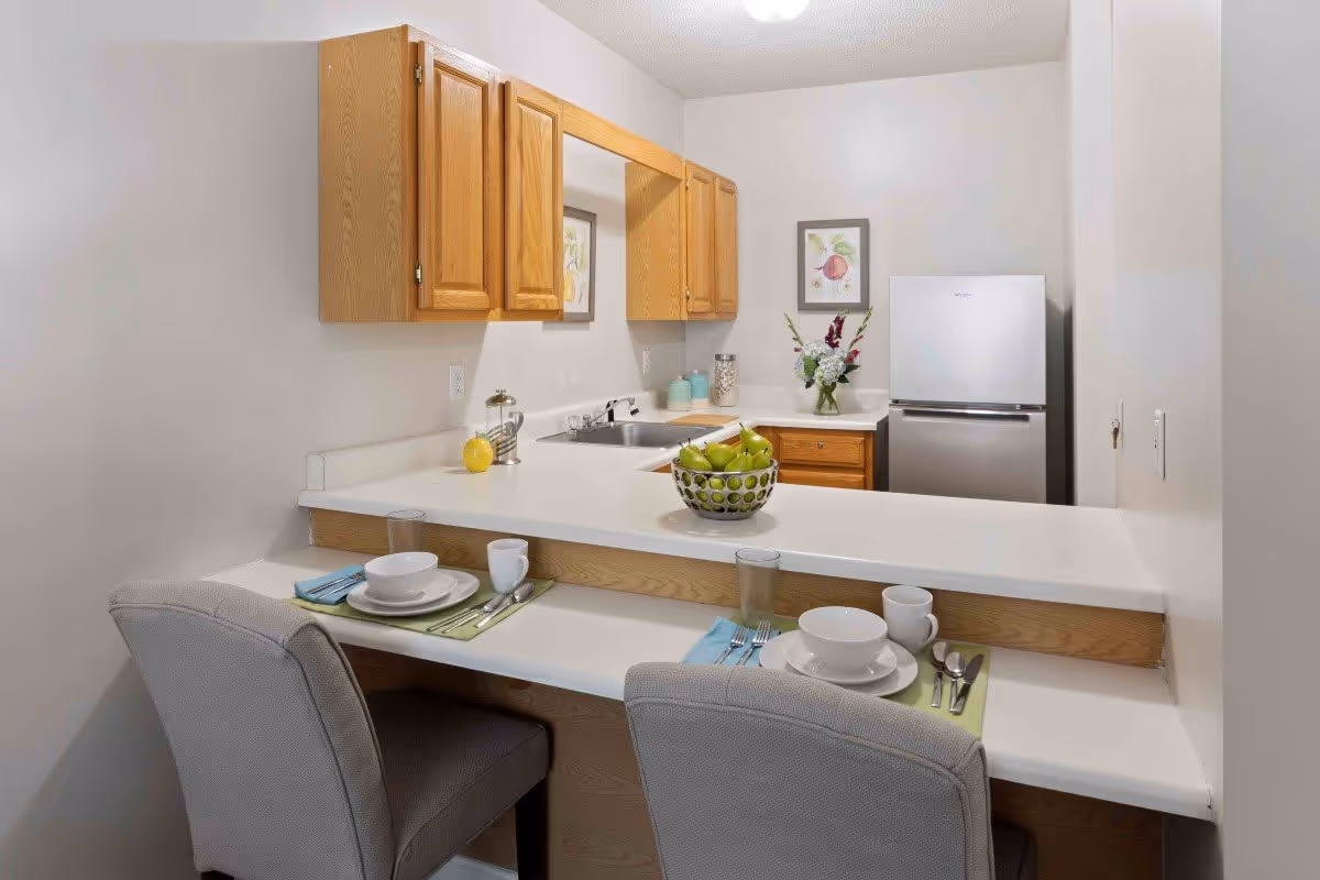 A small kitchen area with wooden cabinets, a stainless steel refrigerator, and a white countertop with a sink. Two place settings with plates, bowls, cups, and utensils are arranged on a breakfast bar with two upholstered chairs. A bowl of green apples and a vase with flowers are on the countertop, and framed artwork hangs on the wall.