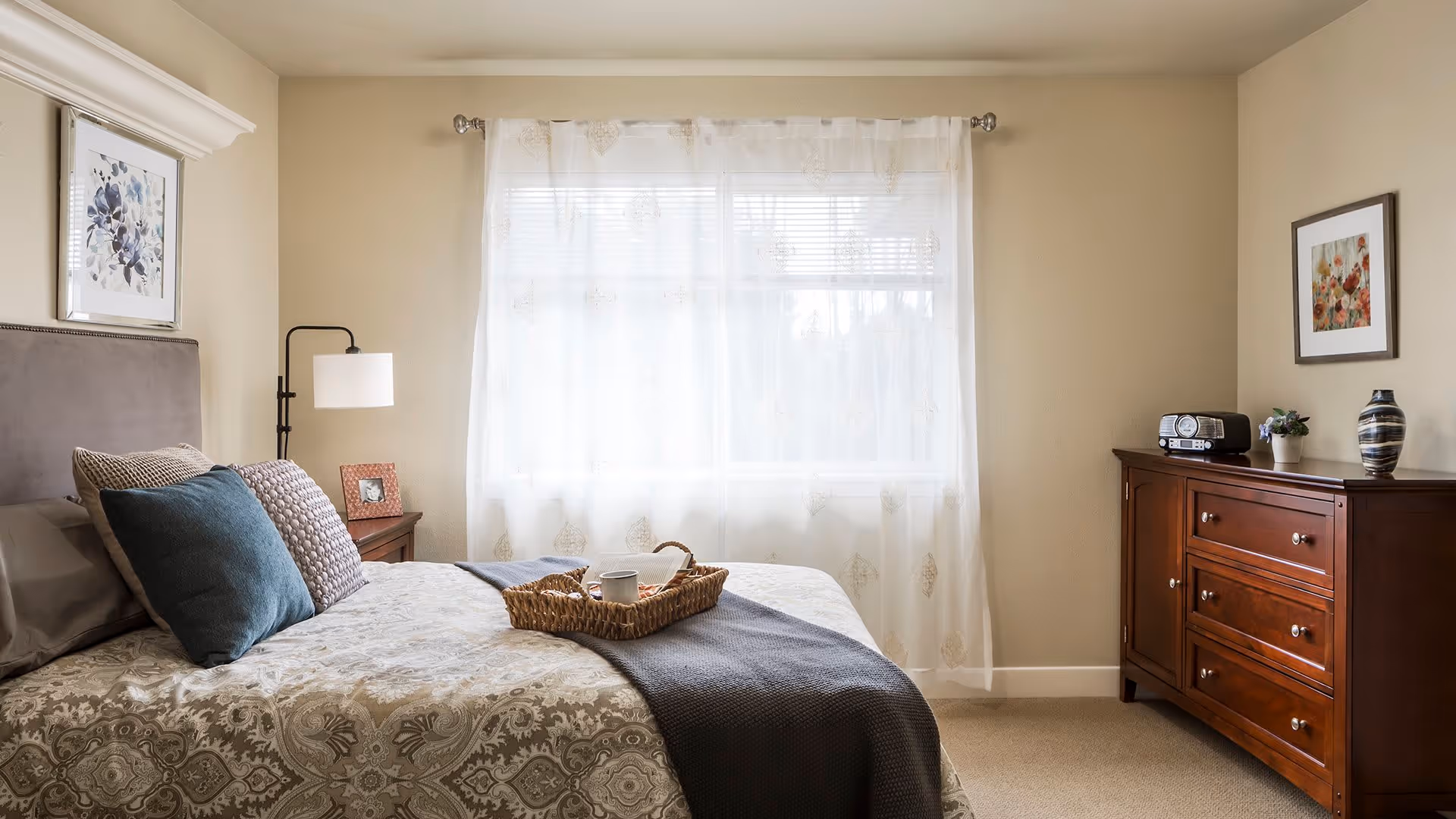 A cozy bedroom with a large window covered by sheer white curtains allowing natural light to fill the room. The bed has a patterned bedspread with multiple pillows and a woven tray holding a cup and a small item. To the left of the bed is a nightstand with a lamp and a framed photo. On the right side of the room is a wooden dresser with a radio, a small plant, and a decorative vase. Two framed artworks hang on the walls.