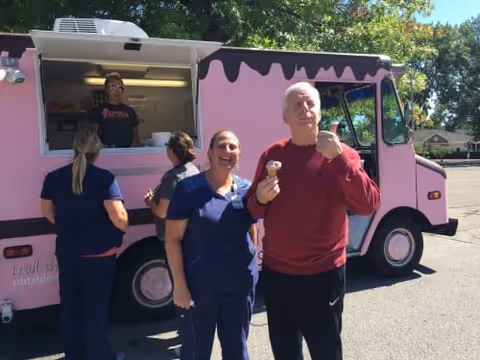 A group of people standing outside a pink food truck on a sunny day. Two women in navy blue uniforms and a man in a red sweater are enjoying ice cream cones, smiling and posing for the photo. Another person is ordering at the food truck window, where a man wearing a dark shirt is serving.