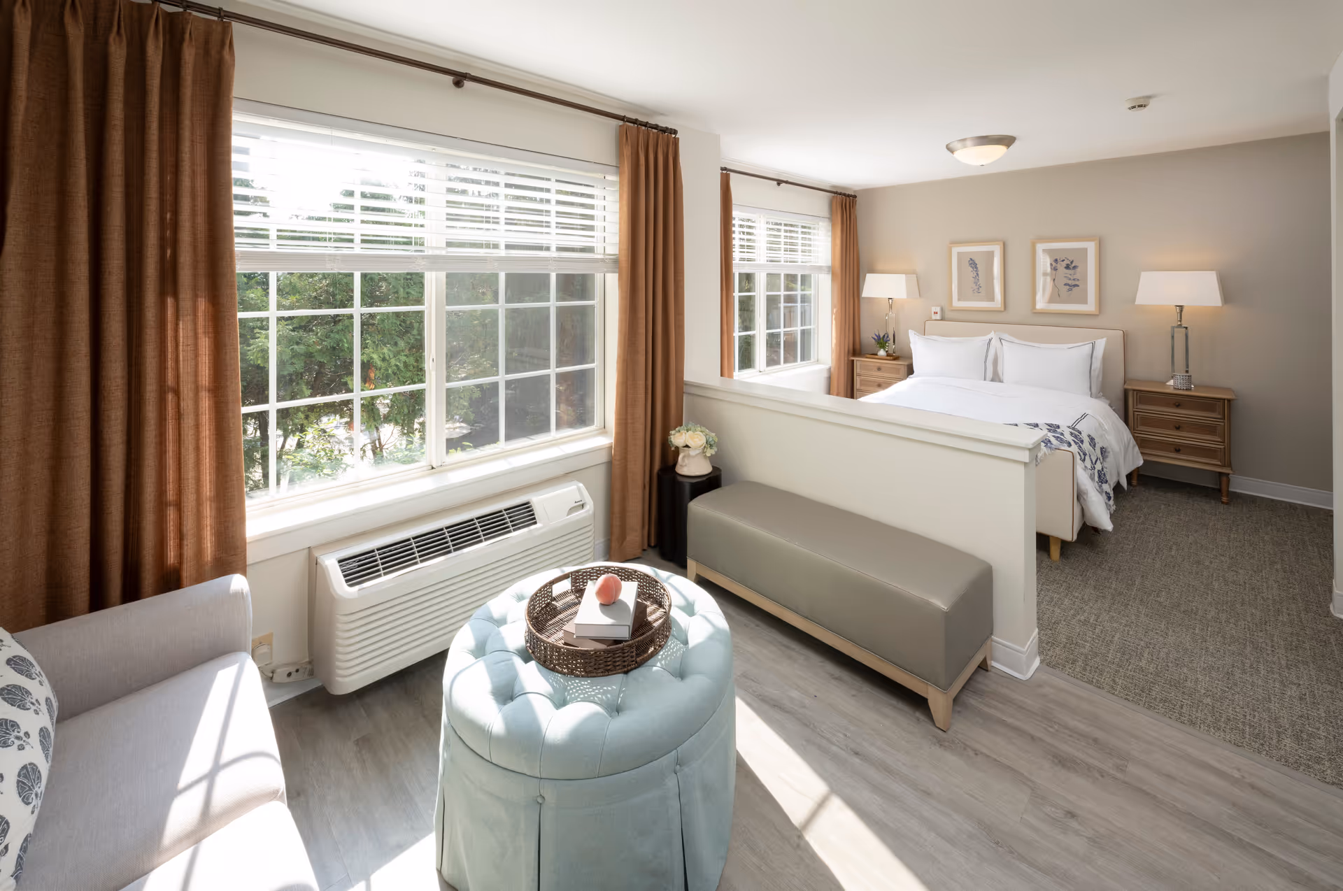 A bright and cozy senior living room and bedroom area with large windows covered by brown curtains and white blinds. The living area has a light gray sofa with a patterned pillow, a round tufted ottoman with a wicker tray holding books and a decorative item, and a gray bench. The bedroom area features a neatly made bed with white linens, two wooden nightstands with lamps, and framed botanical artwork on the wall.
