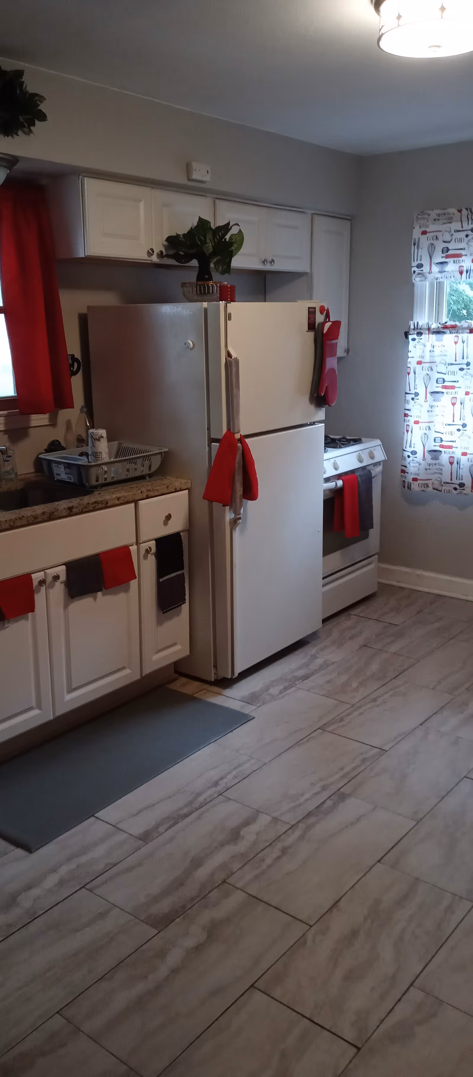 A kitchen with white cabinets, a white refrigerator, and a white stove. The countertop is granite with a dish rack and some items on it. Red and black towels hang on the cabinet handles and the refrigerator door. There is a window with a colorful curtain featuring kitchen utensils, and a red curtain on another window. The floor is tiled with a light gray pattern.