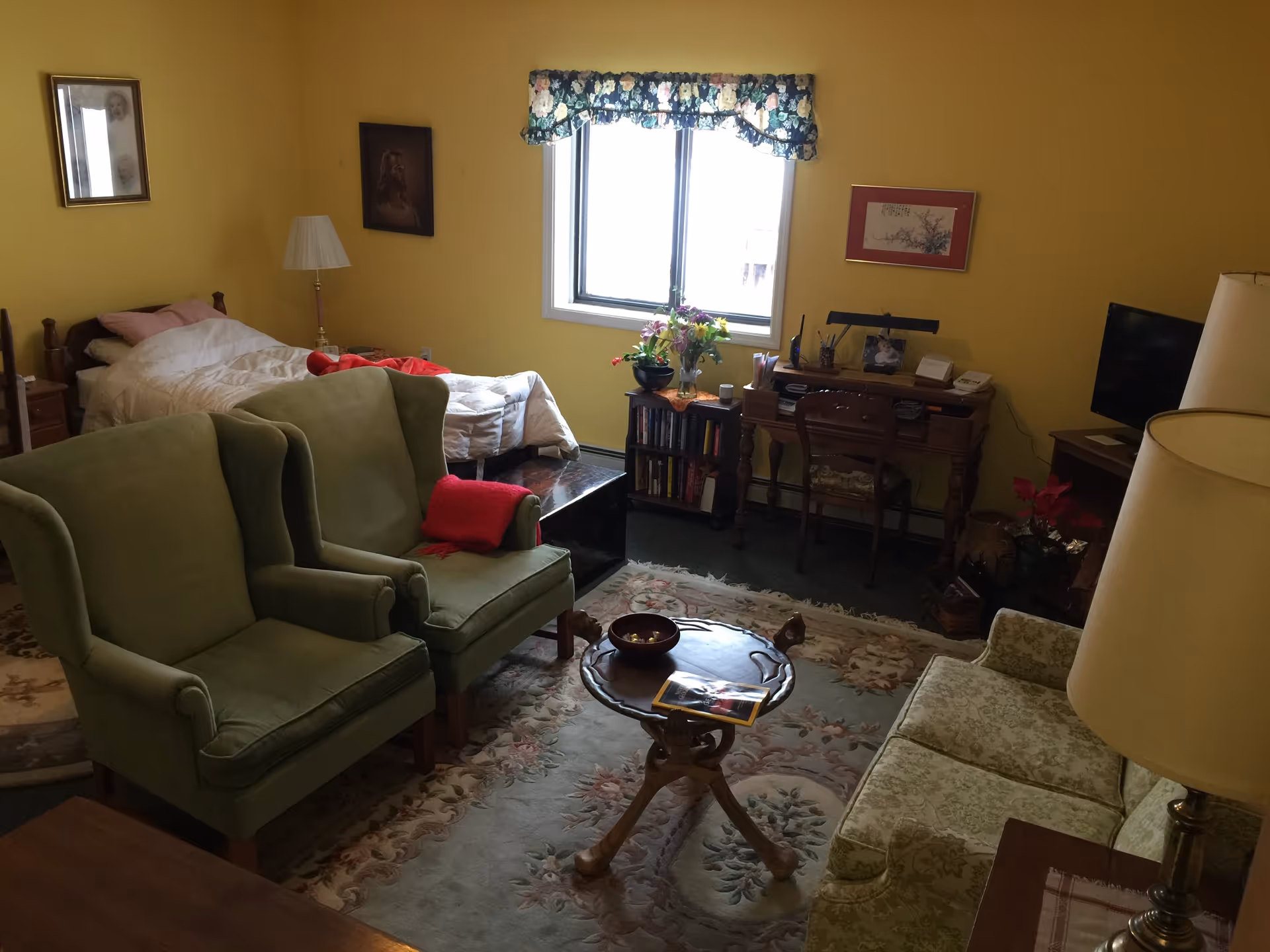 A cozy room with yellow walls featuring a single bed with white bedding and a red blanket, two green upholstered armchairs with a red cushion on one, a small wooden round table, a floral patterned sofa, a desk with a chair, a TV on a stand, and a window with floral valance letting in natural light. The room is decorated with framed pictures, a lamp, flowers, and a bookshelf.