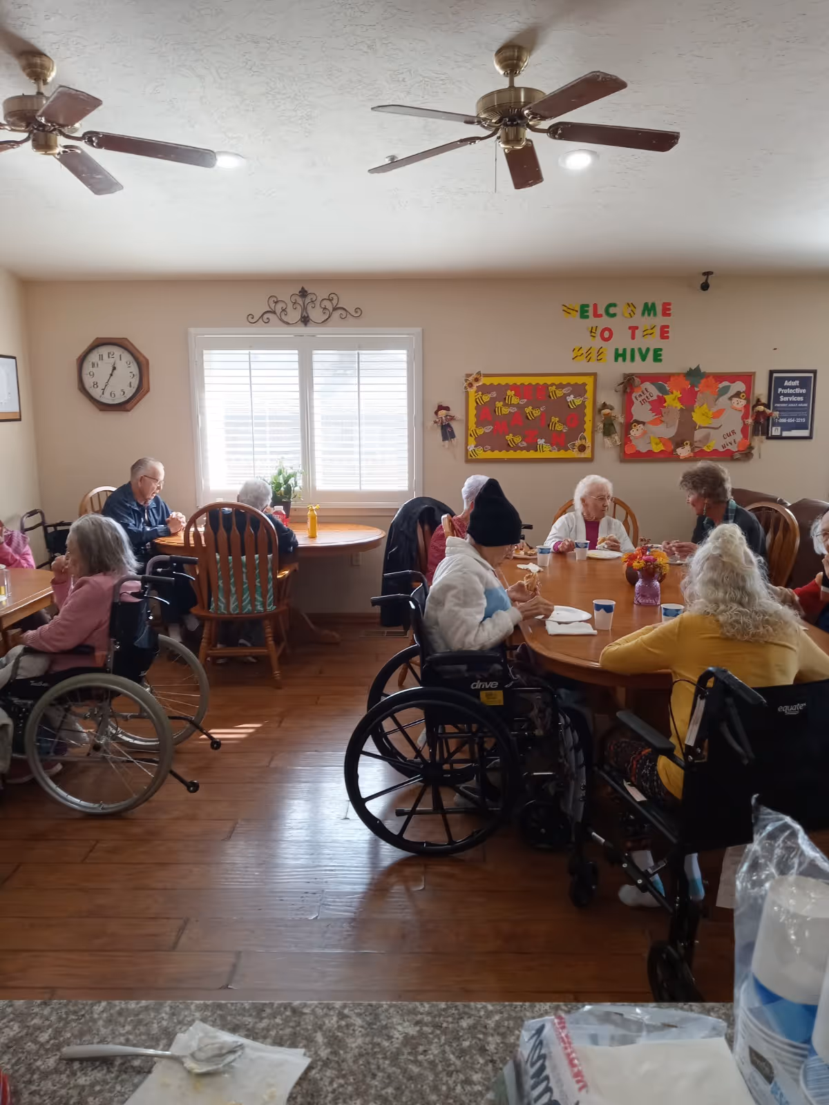 A group of elderly people, some in wheelchairs, sitting around wooden tables in a well-lit room with ceiling fans and a window with blinds. The walls have colorful bulletin boards and a sign that says 'Welcome to the Hive'.